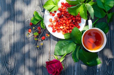 Tahta bir masanın üzerinde yabani olgun çilekler ve bir fincan çay. Terasta güneşli bir günde parlak gölgeler.