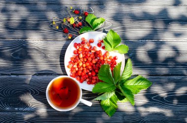 Tahta bir masanın üzerinde yabani olgun çilekler ve bir fincan çay. Terasta güneşli bir günde parlak gölgeler.
