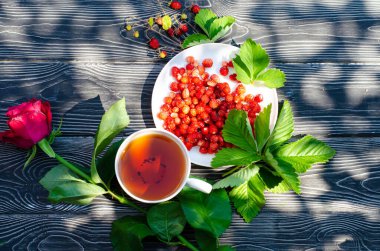 Tahta bir masanın üzerinde yabani olgun çilekler ve bir fincan çay. Terasta güneşli bir günde parlak gölgeler.