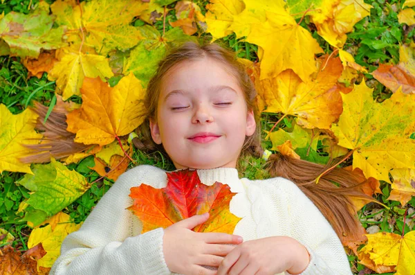 Маленькая девочка лежит на опавших листьях и улыбается. Яркие желтые, оранжевые листья карточек в осеннем парке. — стоковое фото