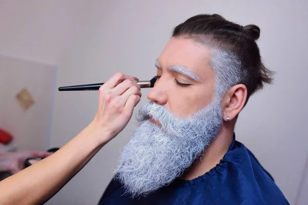 Maquillador hace maquillaje a un hombre con barba gris . — Foto de Stock