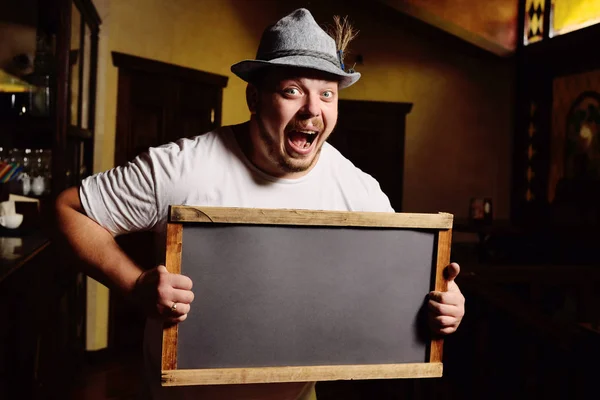 cute cheerful fat man in a Bavarian hat holding a chalkboard or a plate on the background of a pub