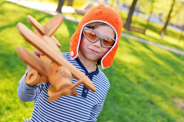Menino em piloto capacete laranja jogando no brinquedo avião de madeira contra fundo grama — Fotografia de Stock