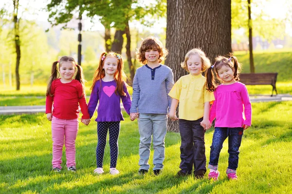 Um grupo de crianças pequenas sorrindo de mãos dadas em um fundo de grama, uma árvore e um parque . — Fotografia de Stock