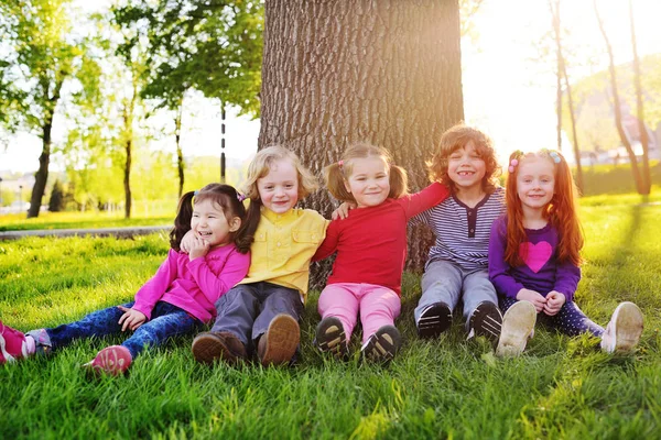 Группа счастливых маленьких детей, улыбающихся сидя в парке на траве под деревом . — стоковое фото