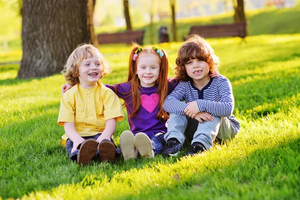 Groep gelukkig weinig kinderen glimlachend in park op gras onder een boom zitten. — Stockfoto