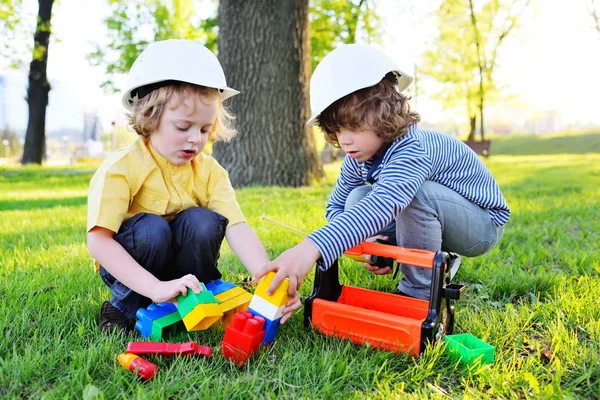 Enfants mignons dans la construction de casques jouent chez les travailleurs ou les constructeurs avec des outils jouets dans un parc sur l'herbe . — Photo