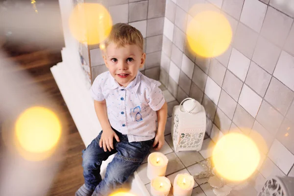 Boy sitting by the fireplace smiles against the Christmas lights background — Stock Photo, Image