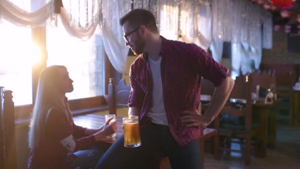 Друзья - молодые парни и девушки пьют пиво, разговаривают и улыбаются в баре — стоковое видео