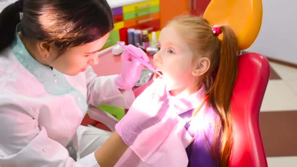 Παιδική οδοντίατρος εξετάζει τα δόντια και το στόμα του ένα χαριτωμένο μικρό κοκκινομάλλα κοπέλα στην οδοντιατρική καρέκλα ένα κίτρινο-κόκκινο. — Αρχείο Βίντεο