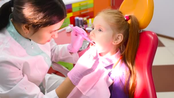 Dentista de niños examina los dientes y la boca de una linda niña de pelo rojo en una silla dental de color amarillo-rojo . — Vídeos de Stock