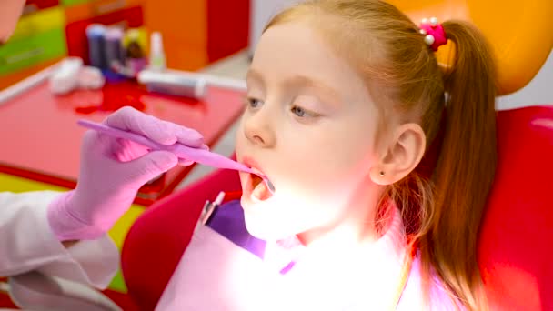 Dentiste enfants examine les dents et la bouche d'une jolie petite fille aux cheveux roux dans une chaise dentaire jaune-rouge . — Video
