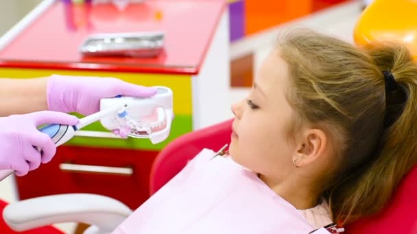 El dentista enseña al niño - una niña linda sin dientes de leche delanteros cómo limpiar los dientes sentados en una silla dental . — Vídeo de stock