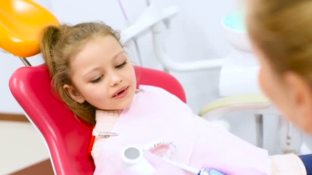 The dentist teaches the child - a little cute girl without front milk teeth how to clean teeth sitting in a dental chair. — Stock Video