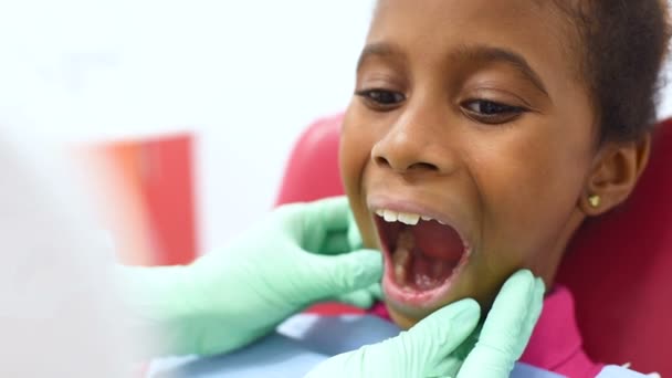 Милая темнокожая афроамериканская девушка сидит в красно-желтом детском стоматологическом кресле у дантиста — стоковое видео
