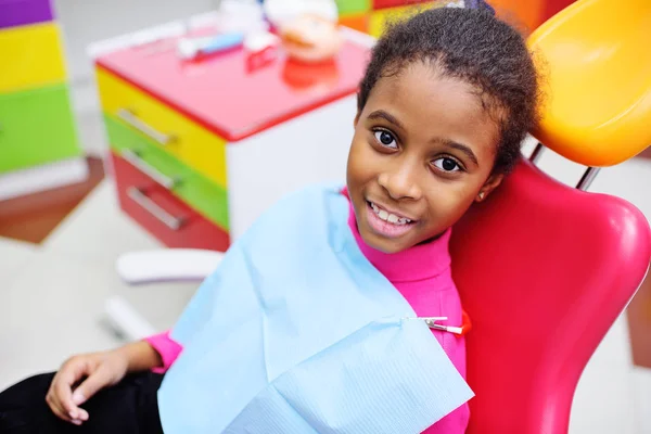 Aranyos fekete kislány mosolyogva ül egy piros fogászati szék — Stock Fotó