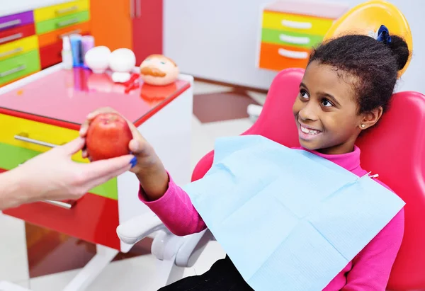 dentist gives a ripe red Apple to a little black African American girl in a dental chair.