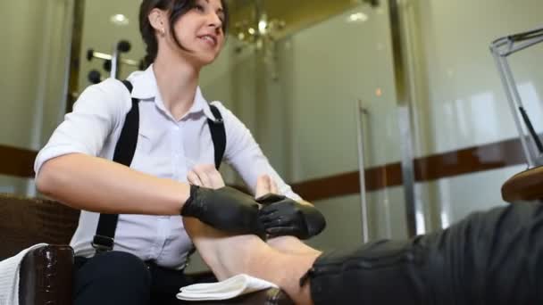 En ung flicka gör en man pedikyr i bakgrunden av en skönhetssalong. — Stockvideo