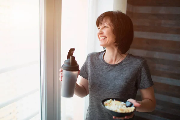 Vuxen passform smal kvinna poserar på kameran. Håller skålen och flaskan i händerna. Äta mat efter träning eller träning. Njuter av maten ensam i rummet. — Stockfoto