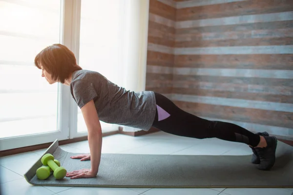 Adulto em forma de mulher magra tem treino em casa. Vista lateral do suporte de modelo sênior bem construído forte em posição de prancha completa. Alongamento para trás e corpo . — Fotografia de Stock