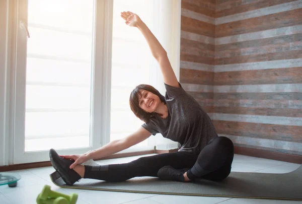 Vuxen passform smal kvinna har träning hemma. Hon gör stretching på en yogamatta vid fönstret Ta hand om kroppen — Stockfoto