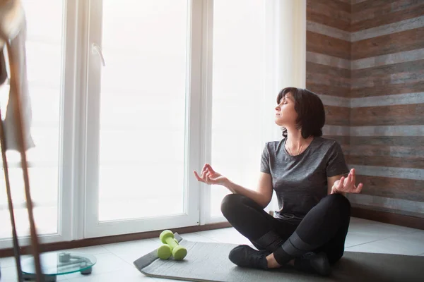 Vuxen passform smal kvinna har träning hemma. Processen för äldre modell meditera. Håll händerna på knä och titta på fönstret. — Stockfoto
