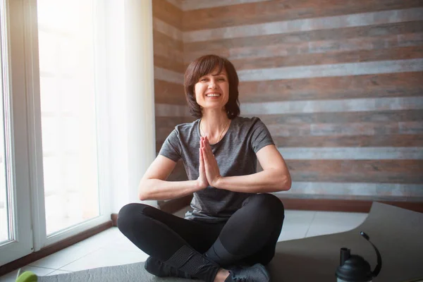 Vuxen passform smal kvinna har träning hemma. Glada positiva kvinnliga person hålla händerna tillsammans i be positiob och titta på kameran med leende. Meditera eller asana pose. — Stockfoto