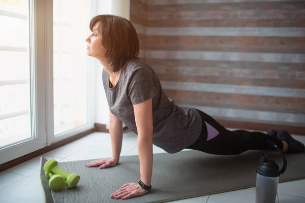 Vuxen passform smal kvinna har träning hemma. Allvarlig mogen kvinnlig person står på yogapositionen för att få styrka och välbefinnande. Ensam övning. — Stockfoto