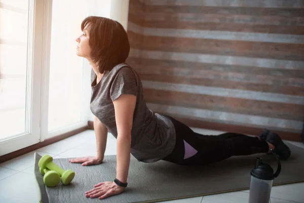 Vuxen passform smal kvinna har träning hemma. Sidovy av senior modell stående på knä och hålla kroppen på mattan. Stretching kroppen träning och träning. — Stockfoto