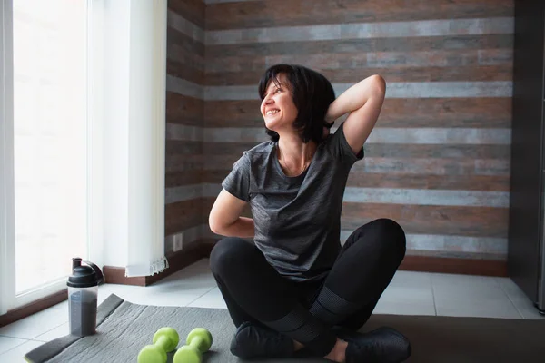 Vuxen passform smal kvinna har träning hemma. Fantastiskt positiv kvinnlig mogen person hålla händerna tillsammans på baksidan och titta med leende på fönstret. stretcharmar och träning. — Stockfoto