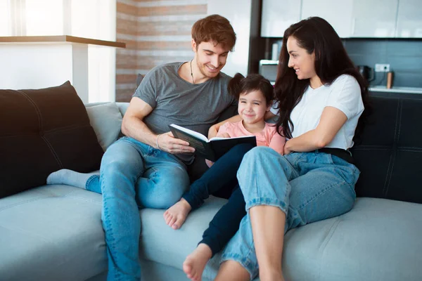 Rodina doma čte knížku. Dospělý bratr a sestra čtou knížku malé sestře. Mladí rodiče a dcera — Stock fotografie
