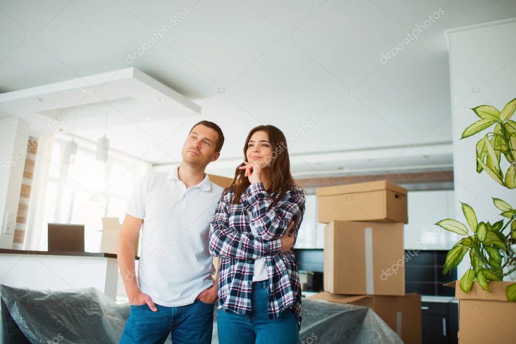 A young married couple in the living room in the house stand near unpacked boxes. hey are happy about new home. Moving, buying a house, apartment concept.