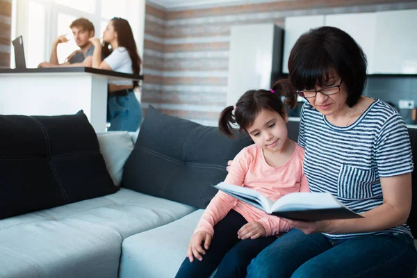 Grandmother reads a book to her granddaughter. Father and mother use laptop on background. Big family at home. — Stock Photo, Image