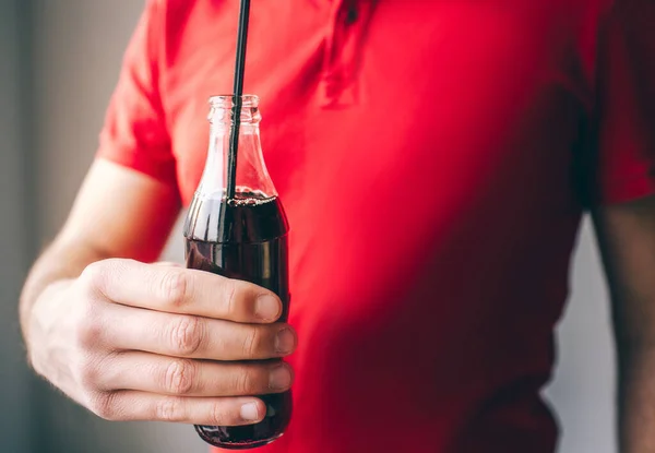 Hombre joven con camisa roja aislado sobre el fondo. Guy sostiene la botella de vidrio en la mano con paja de plástico en el interior. Coca cola deliciosa agua carbonatada . — Foto de Stock