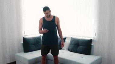 Genç adam evde egzersiz yapıyor. Adam akıllı telefonuyla kulaklıkla müzik açıyor ve oturma odasında aynı yerde koşmaya başlıyor. Egzersizden önce bacakları ısıtıyorum..