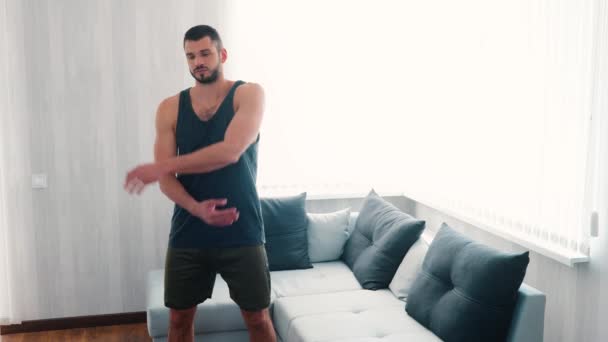 Un joven haciendo ejercicio en casa. Fuerte poderoso culturista no profesional estirando las manos y la parte superior del cuerpo antes del entrenamiento. Hacer ejercicio solo en la sala de estar . — Vídeo de stock