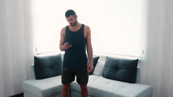 Un joven haciendo ejercicio en casa. cámara lenta de chico encendiendo música en los auriculares con el teléfono inteligente y empezar a funcionar en el mismo lugar en la habitación. Calentar el cuerpo antes de entrenar y hacer ejercicio . — Vídeos de Stock