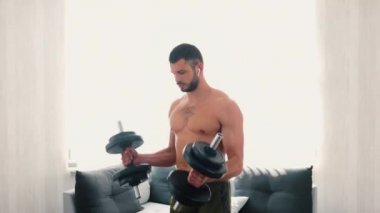 Genç adam evde egzersiz yapıyor. Kas geliştirici oturma odasında egzersiz yapıyor. Büyük, ağır dambılları tutarak ve kaldırarak. Güçlü bir eğitim..