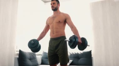 Genç adam evde egzersiz yapıyor. Dambıllarla egzersiz yaparken kameranın yavaşça geriye doğru hareket etmesi. Ellerinizi kaldırın ve kaslarınızı çalıştırın..