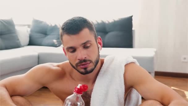 Junger Mann übt zu Hause. Kamera bewegt sich rückwärts. Mann trinkt Wasser und schaut auf Smartphone in der Hand. Ruhe nach dem Training oder Training. Suche im Internet. — Stockvideo