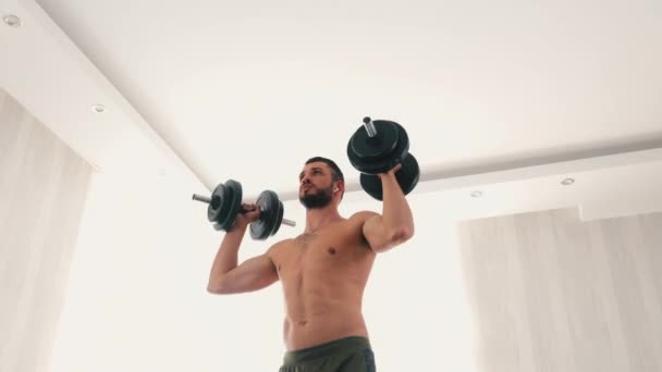 若い男が家で運動している。リビングルームでの強いセクシーな強力な男のトレーニングボディの低ビュー。手に重いダンベルを保持し、それらを上げる。パワートレーニング. — ストック動画