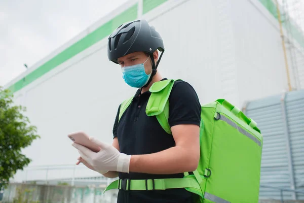 Food delivery concept. The food delivery man uses a smartphone to reach customers faster. Courier has a refrigerator in a green backpack. He is wearing a medical mask nd gloves