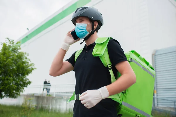 Food delivery concept. The food delivery man uses a smartphone to reach customers faster. Courier has a refrigerator in a green backpack. He is wearing a medical mask nd gloves