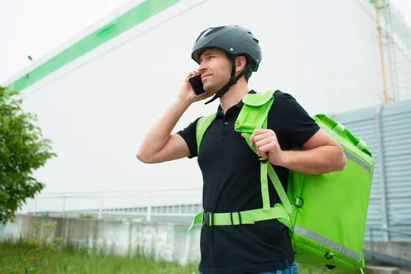 Food delivery concept. The food delivery man uses a smartphone to reach customers faster. Courier has a refrigerator in a green backpack.