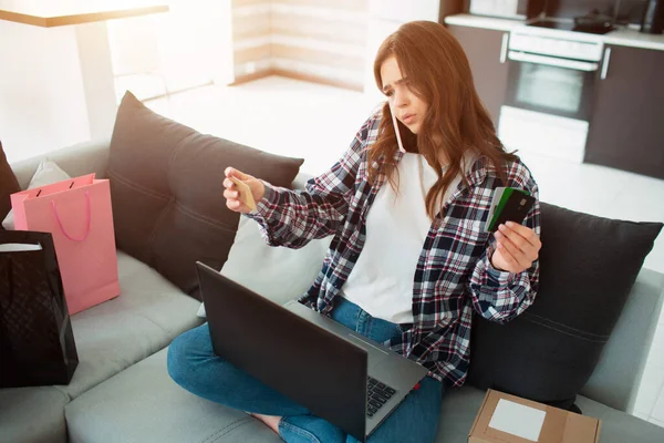 Compras en línea. Una mujer joven se sienta en un sofá y dicta los datos de su tarjeta de crédito a un vendedor en una tienda en línea . — Foto de Stock