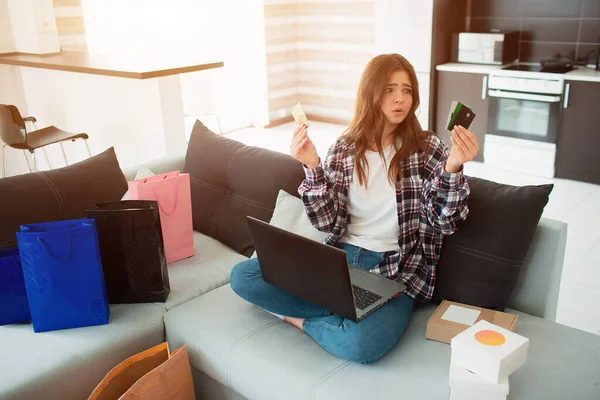 Compras en línea y entrega de bienes o compras a casa. Una mujer joven se sienta en casa en el sofá y no sabe qué tarjeta de crédito o débito el banco tiene que elegir para comprar bienes o cosas . — Foto de Stock