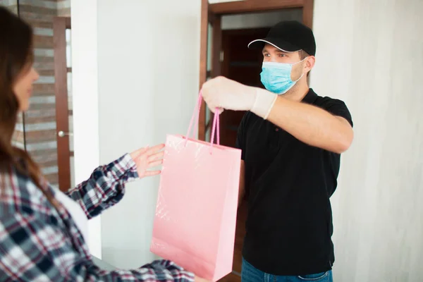 Delivery of goods and purchases home. The courier or delivery man passes the package with the goods to a young woman. She pays for his work with a credit card. He is wearing a mask