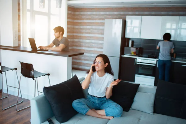 Familia en casa. Cada uno está ocupado con sus propios asuntos. Una joven está hablando por teléfono. Un hombre usa un portátil mientras una mujer adulta cocina en la cocina — Foto de Stock