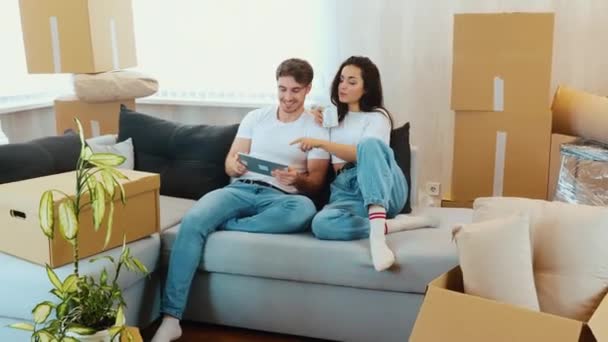 Junges Paar bezieht neue Wohnung Mann und Frau sitzen zusammen auf dem Sofa. Guy hält Tablette und Hobel. Mädchen trinken aus weißer Tasse. Mitten im Umzugsprozess. — Stockvideo