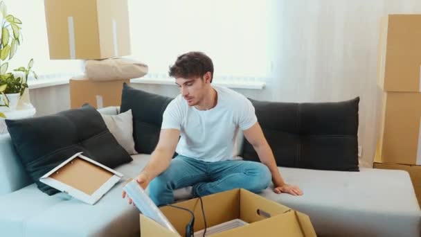 Un joven se muda a un apartamento nuevo. Siéntese en el sofá solo y desempaque o desempaque el envase de cartón. Saca el marco y otras cosas. Tipo cansado cayendo de nuevo en el sofá . — Vídeos de Stock